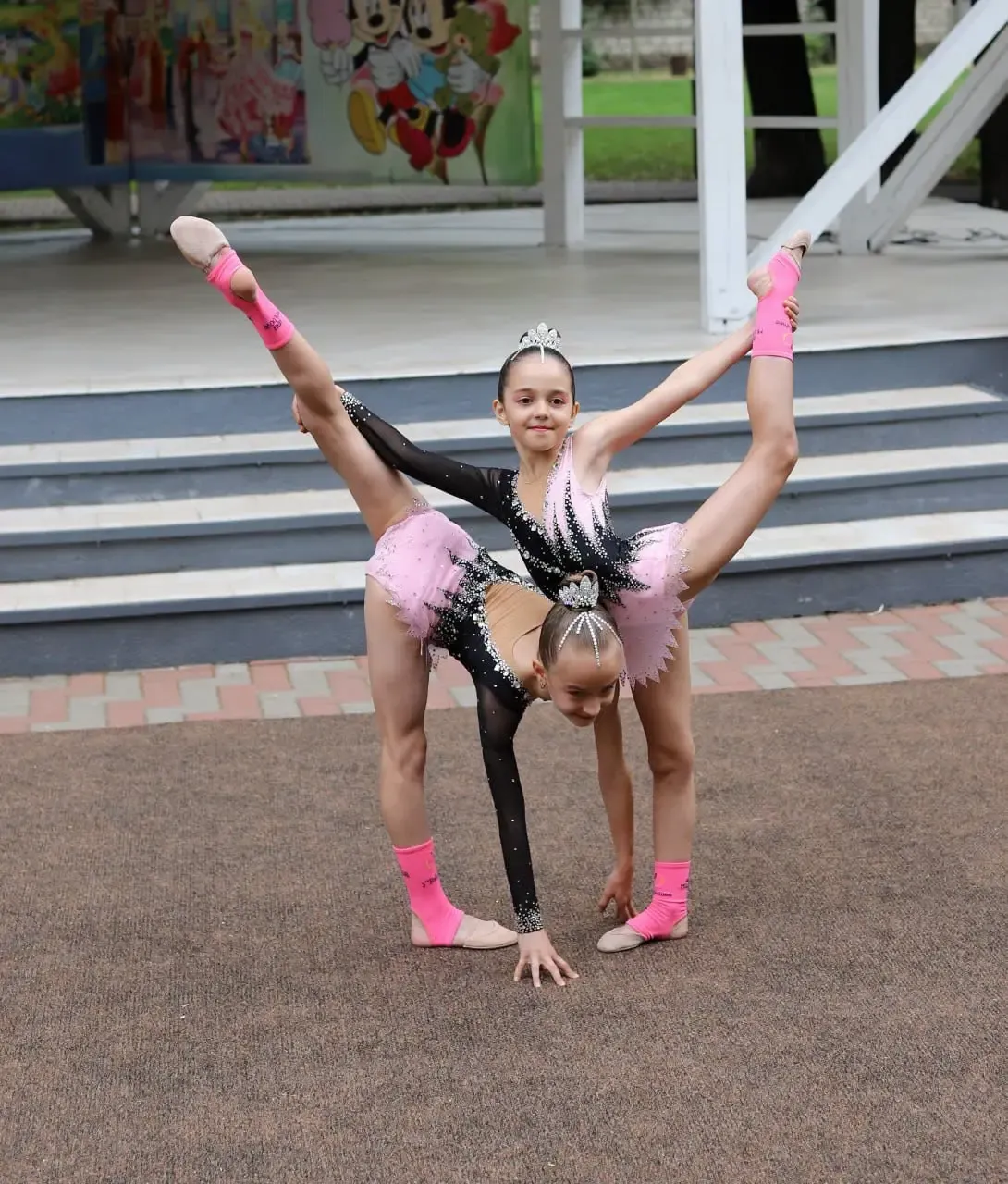 Două gimnaste execută un număr de acrobatică pe scena din parc: una, în costum negru cu paiete, ține o cumpănă cu piciorul întins vertical, în timp ce a doua, în costum roz, face un pod dedesubt.