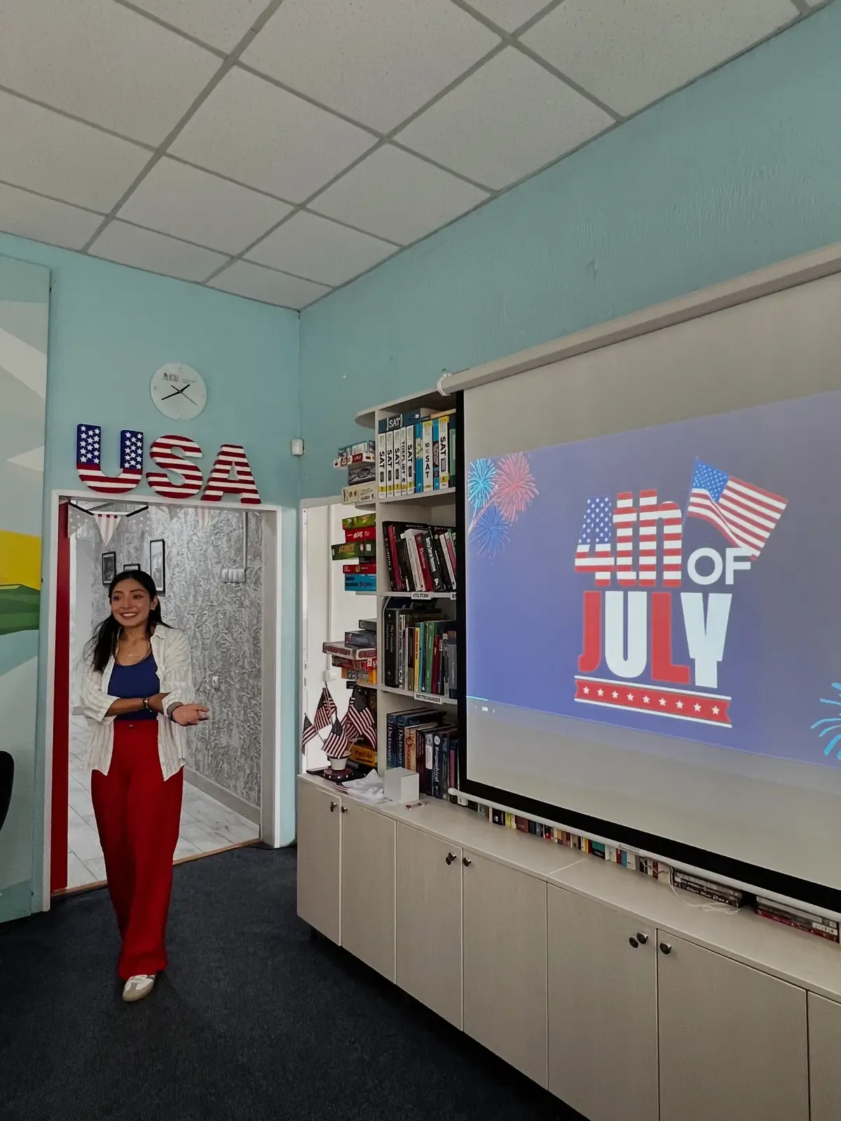 Interiorul American Corner Ungheni: literele decorative „USA" în roșu-alb-albastru deasupra unei uși, un ecran de proiecție cu mesajul „4th of July" și rafturi cu manuale SAT și TOEFL și cărți în limba engleză.