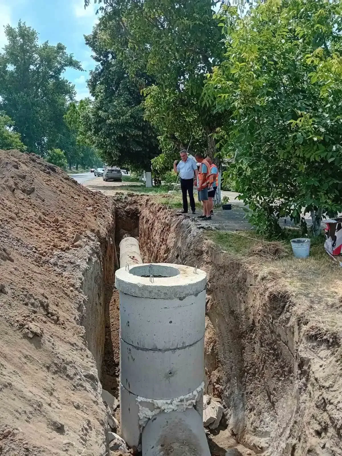 Un cămin de beton pentru canalizarea pluvială, montat într-o tranșee săpată pe marginea străzii Decebal, cu o conductă vizibilă în șanț; în fundal, drumul și un muncitor discutând cu un bărbat în cămașă albă.