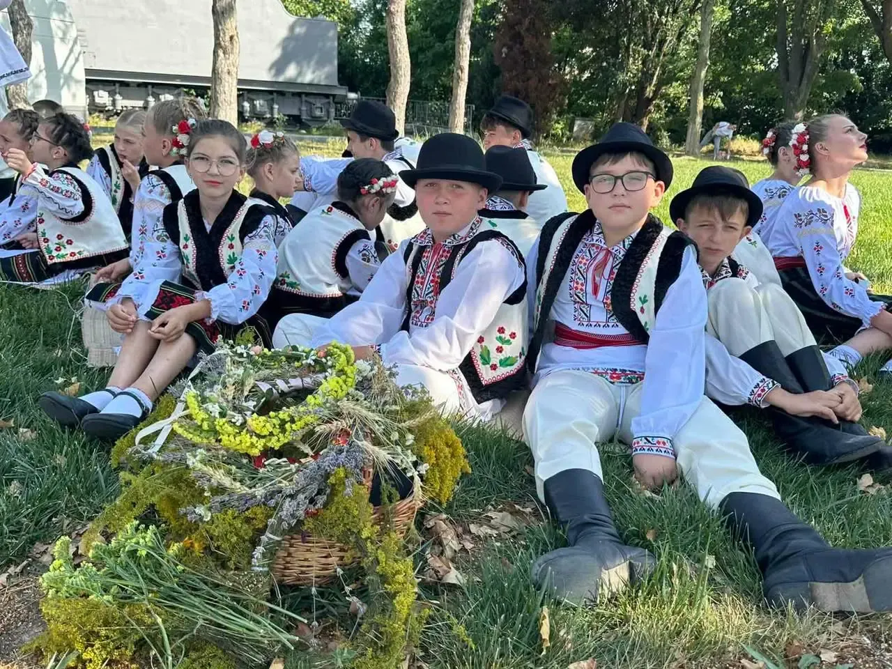 Copii îmbrăcați în costume populare, cu bărbați purtând pălării și fete cu coronițe de flori, așteaptă pe iarbă lângă un coș cu cununi de sânziene și buchete de lavandă.