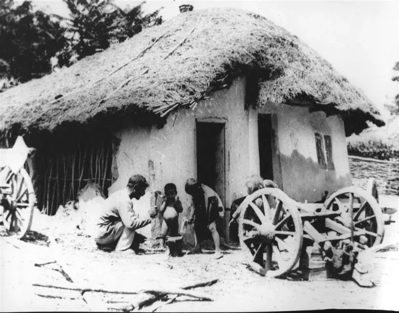 Fotografie alb-negru din 1931: un bărbat ghemuit cu câțiva copii desculți în fața unei case de lut cu acoperiș de stuf, lângă un car cu roți de lemn.