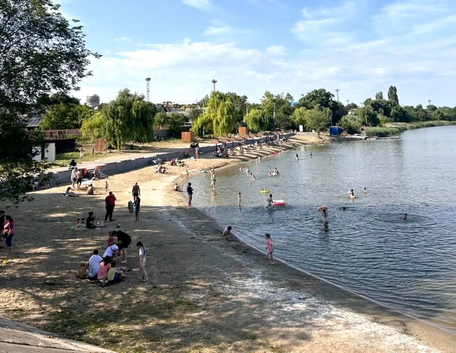 A crowded beach on the shore of a lake: dozens of people, adults and children, on the sand and in the water, on a sunny summer day.