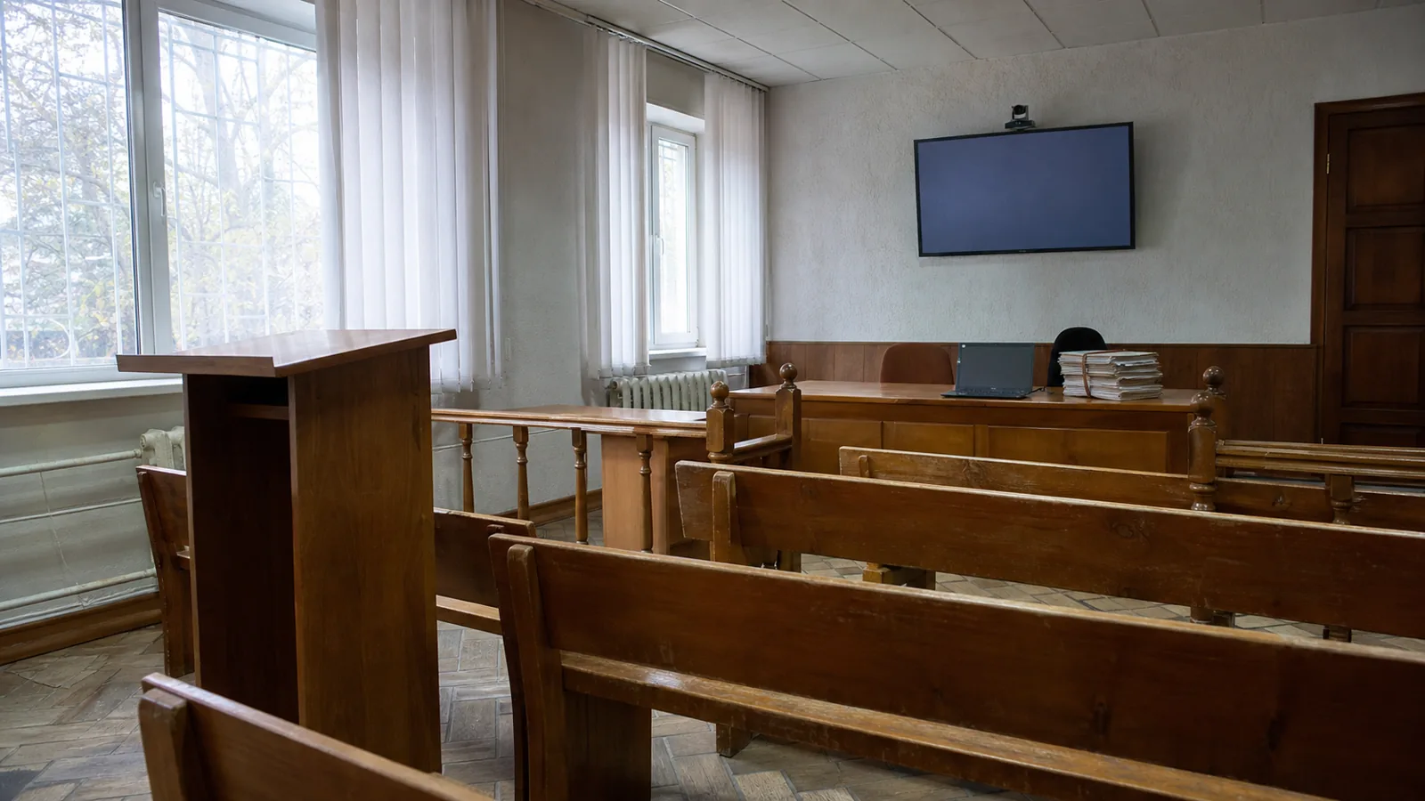 Interiorul unei săli de judecată mici și goale, cu bănci de lemn, un ecran de videoconferință pe perete și un laptop deschis lângă un teanc de dosare legate cu șnur.