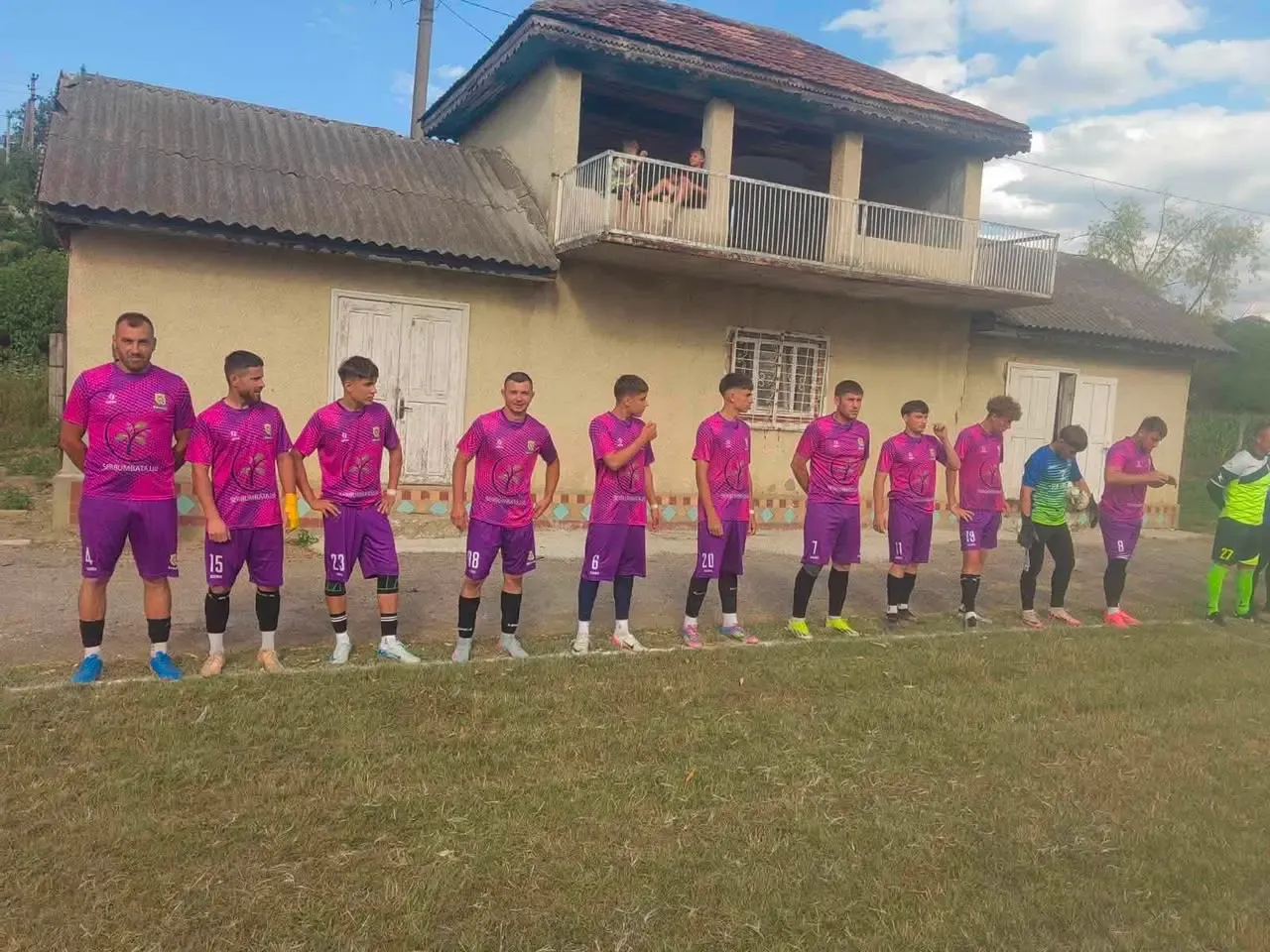 Jucătorii echipei FC Haiduc Bumbăta, în echipament roz cu mov, aliniați pe marginea terenului în fața unei case de sat, înaintea meciului.