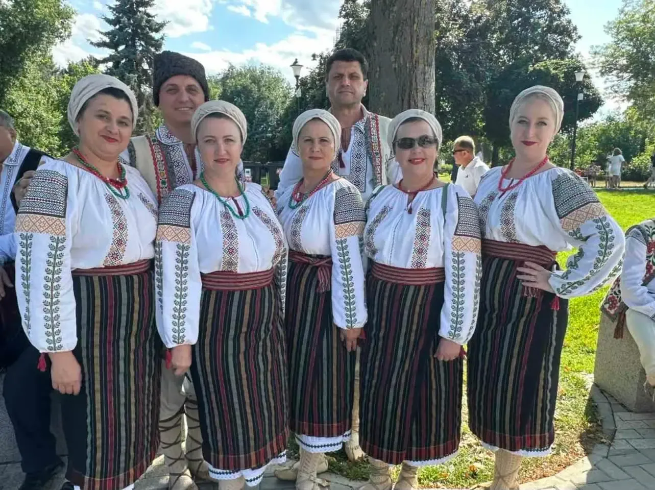 O formație folclorică de adulți, femei în ii și fote cu dungi, cu mărgele de coral, și bărbați în cămăși brodate și căciuli, pozează în grup într-un parc din centrul orașului.