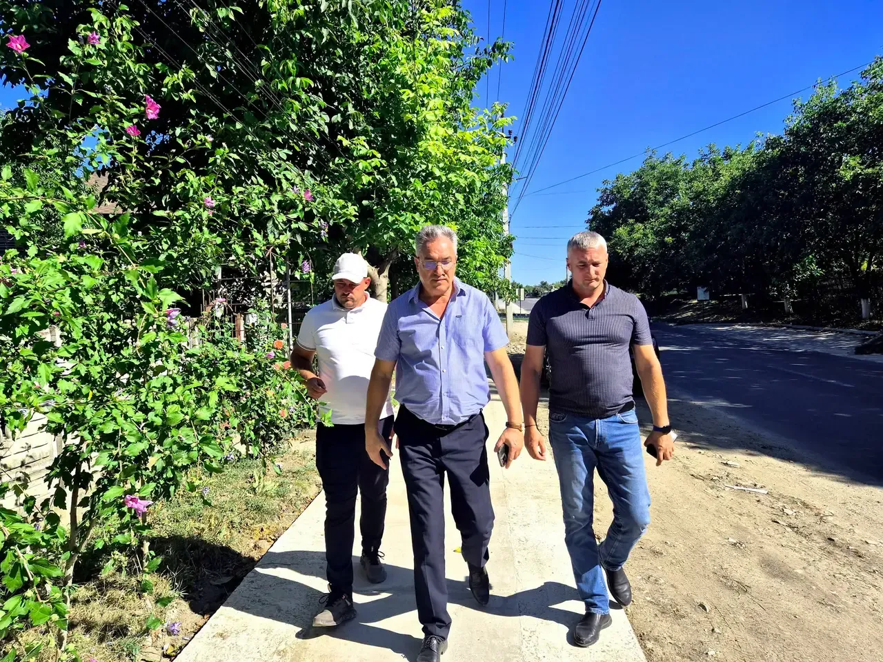 Primarul Vitalie Vrabie merge pe noul trotuar pavat de pe strada Ștefan cel Mare, alături de doi bărbați, cu o stradă asfaltată în dreapta și verdeață pe margine.