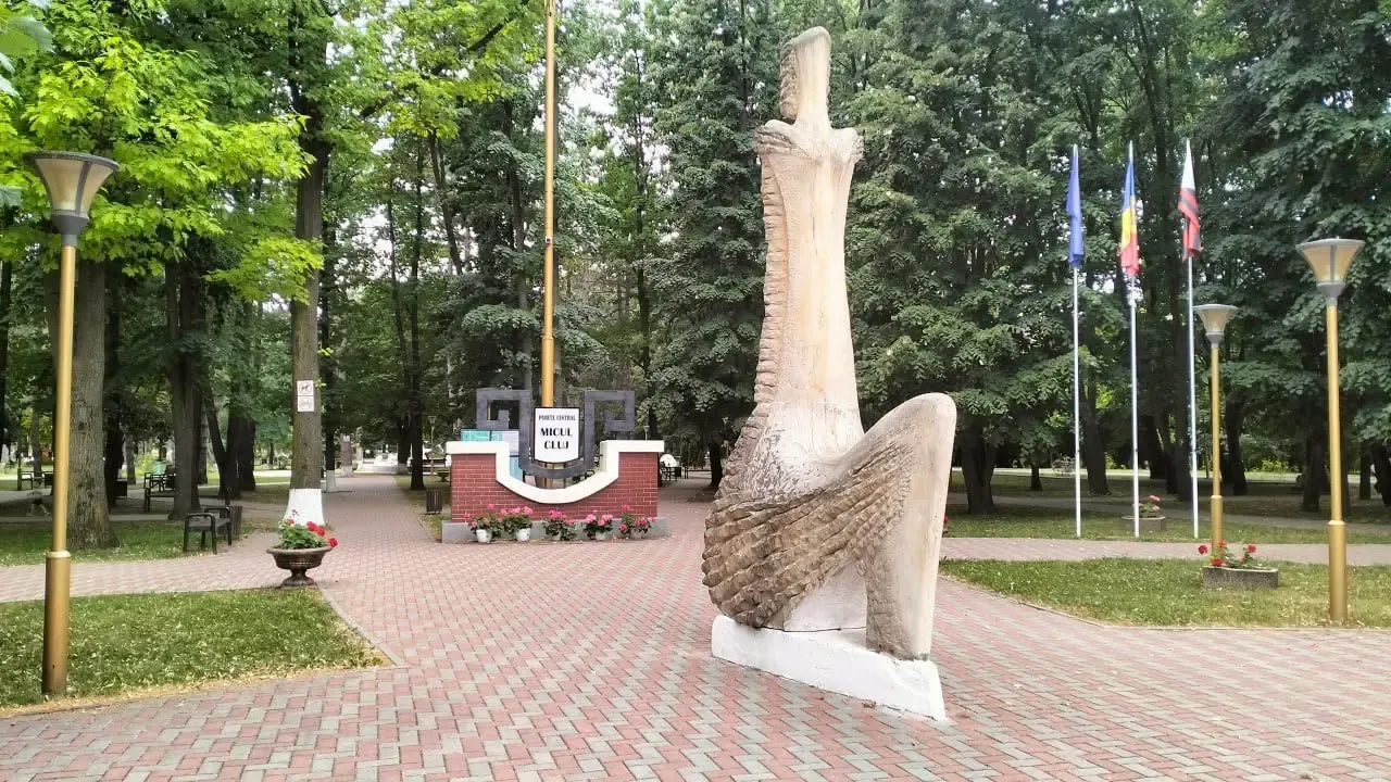 Monumentul „Micul Cluj” de la intrarea în parc — o sculptură albă lângă catargele cu drapelele Moldovei, României și Uniunii Europene, pe o alee pavată dintre brazi.