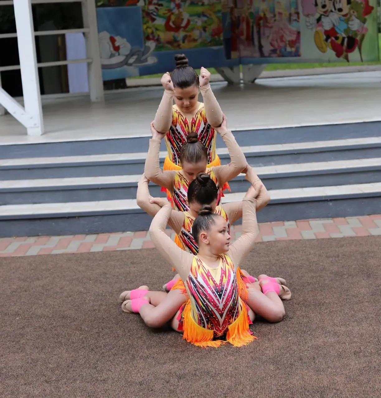 Patru gimnaste în costume portocalii construiesc o piramidă umană pe scenă, una peste alta, pe fundalul murei colorate cu personaje de desene animate.