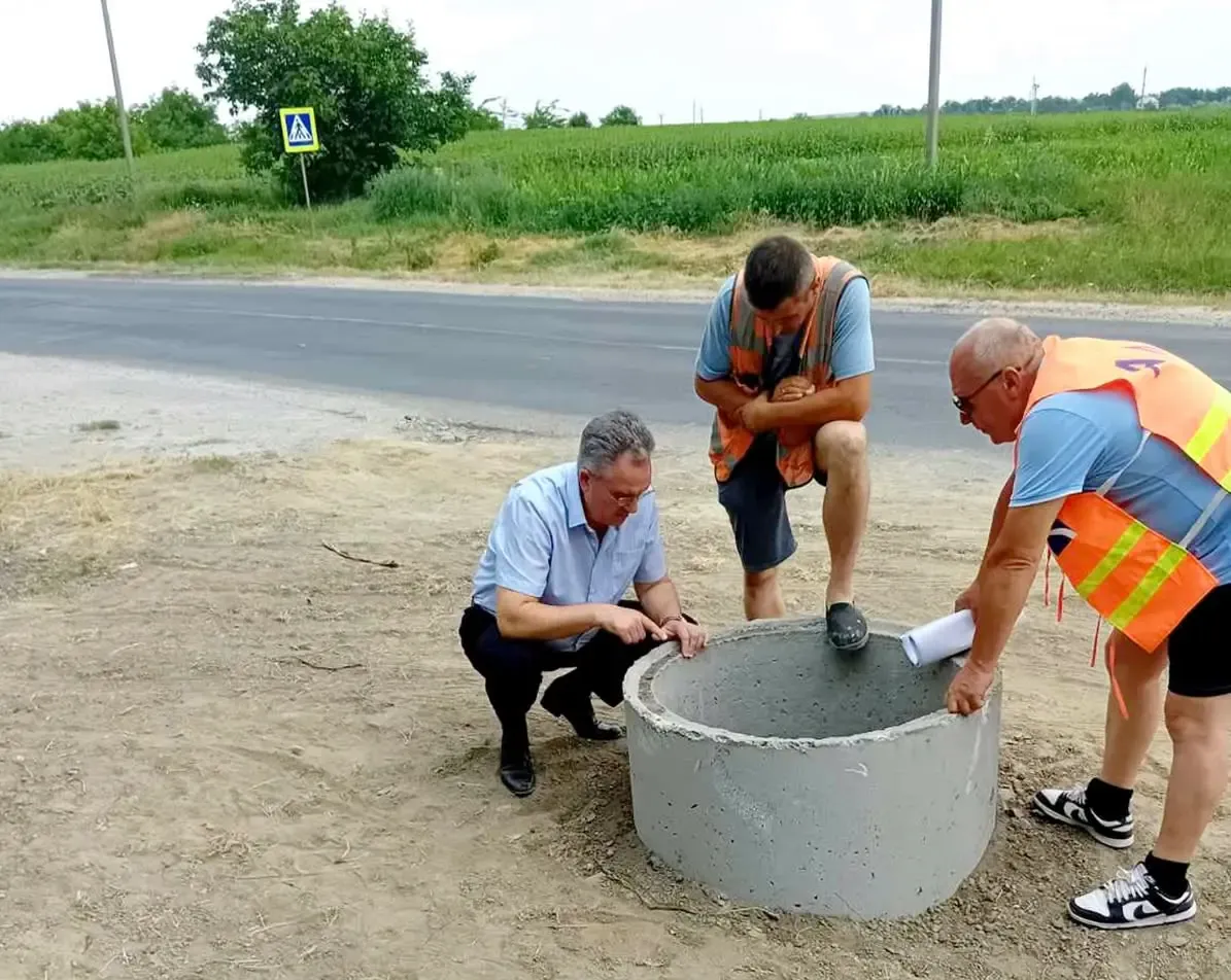 Primarul Vitalie Vrabie, ghemuit lângă un inel de beton pentru cămin, examinează materialele împreună cu doi muncitori în veste reflectorizante, la marginea străzii Decebal.