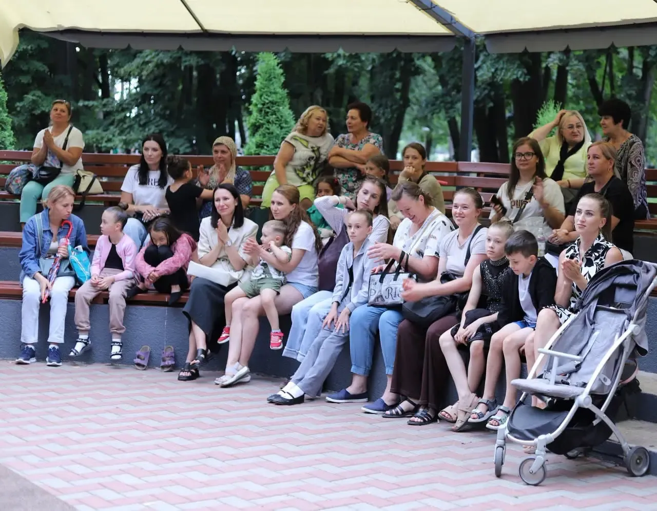 Public numeros pe băncile din parc — mame cu copii, printre care un cărucior — urmărește spectacolul de pe scena din Parcul „Micul Cluj”.