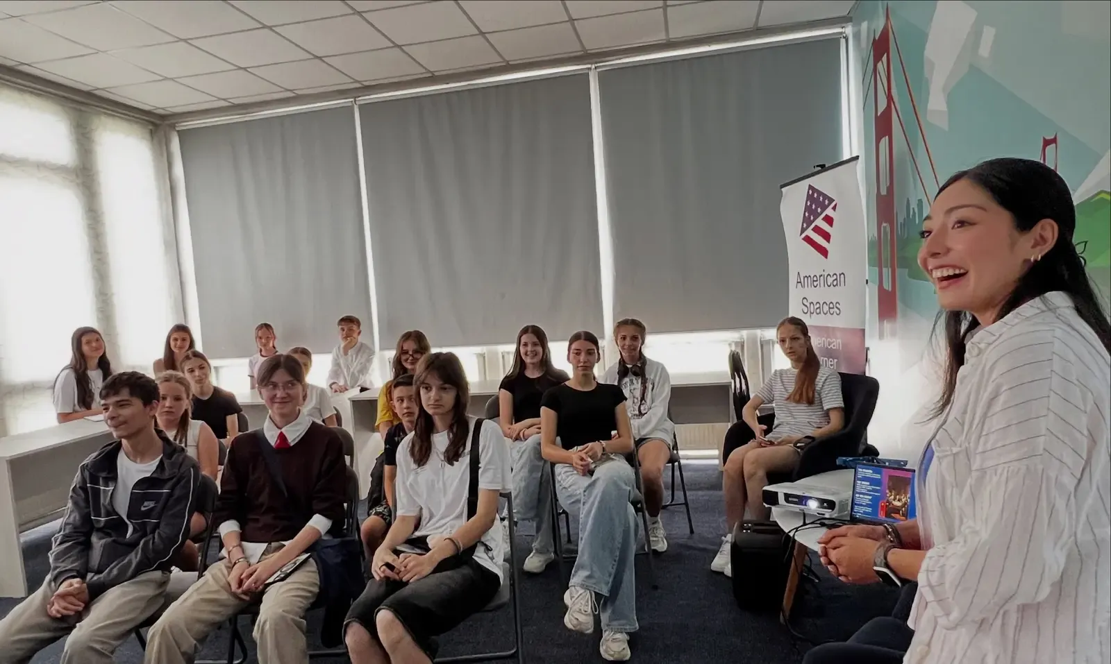 O facilitatoare a American Corner Ungheni conduce un joc interactiv în fața unor tineri ungheneni, la recepția „Freedom 250" de Ziua Independenței SUA; în dreapta, roll-up-ul „American Spaces" și o murală cu podul Golden Gate.