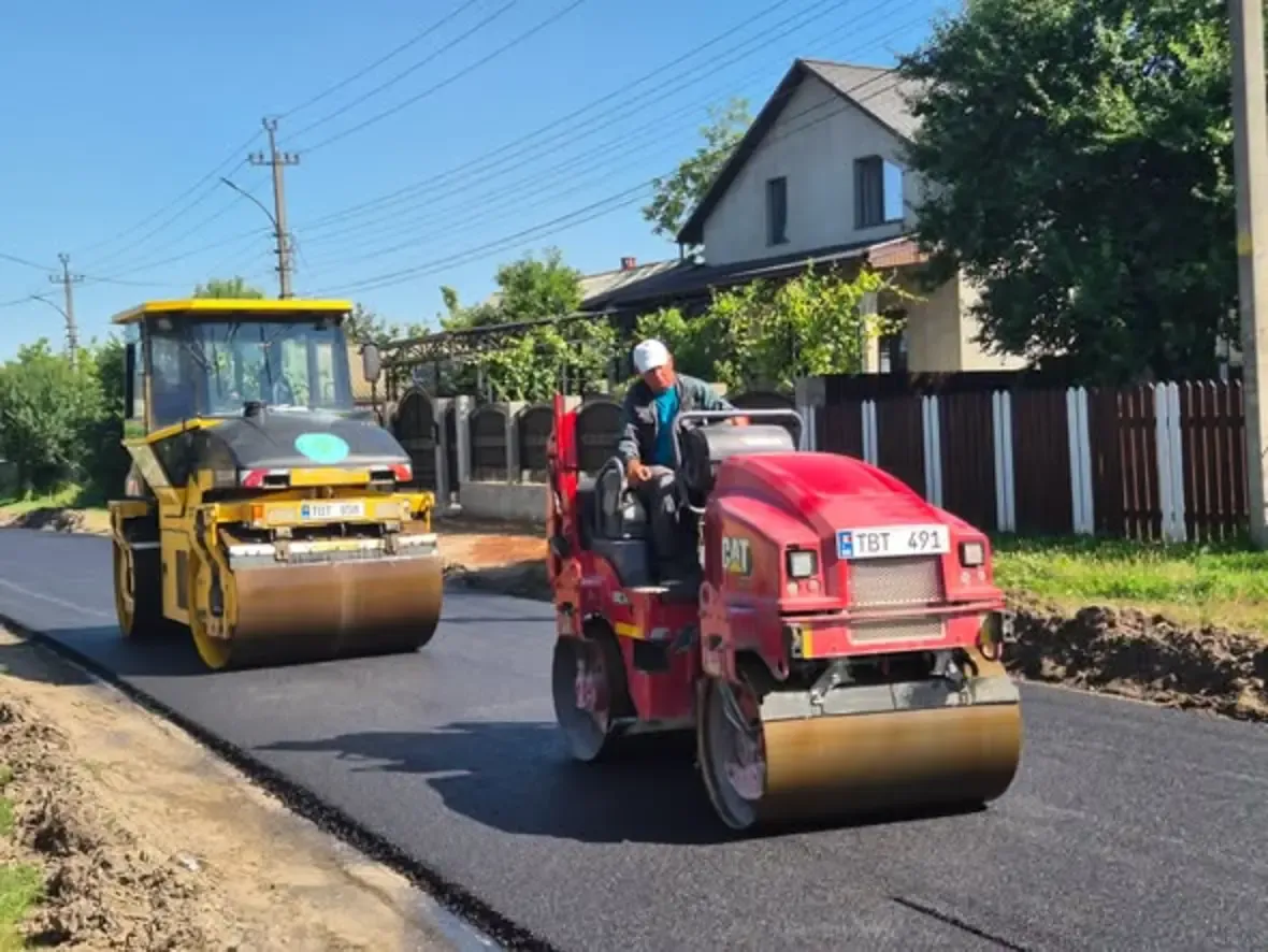 Două cilindre compactoare, unul galben și unul roșu, netezesc stratul proaspăt de asfalt pe o stradă din Ungheni, mărginită de case și garduri.