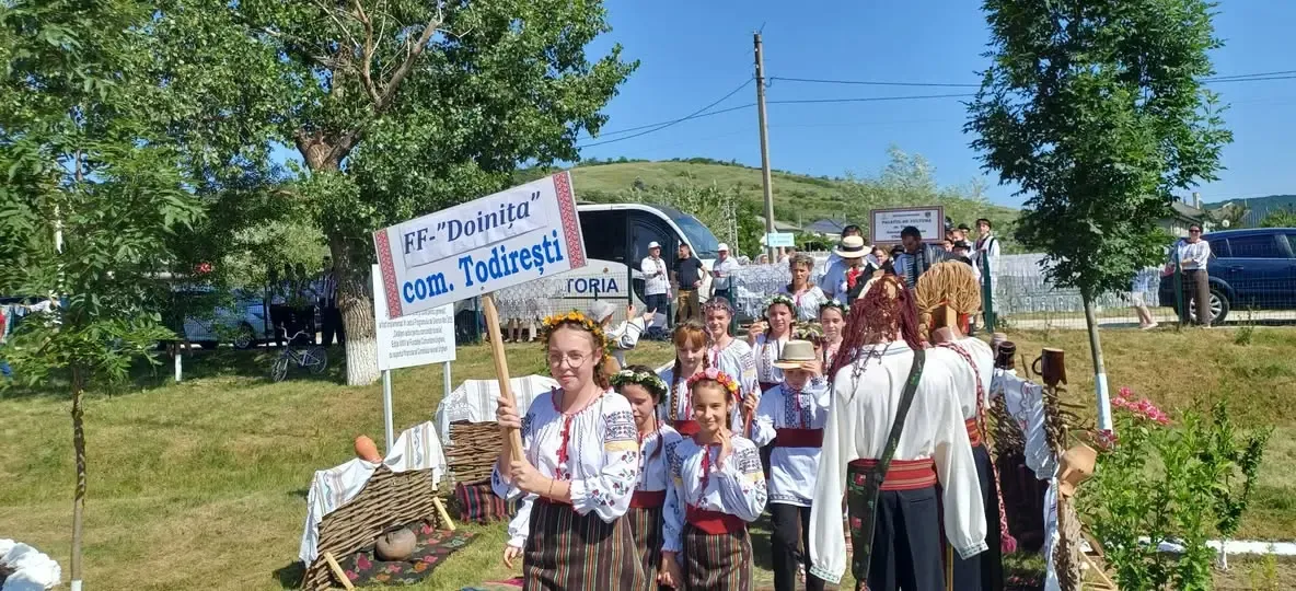 Tineri în costume populare, cu cununi de flori, purtând pancarta unei formații folclorice din comuna Todirești, la „Sărbătoarea Portului Popular” de la Mănoilești.
