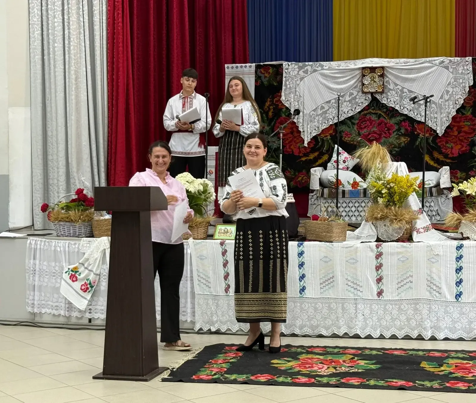Masa de sărbătoare a festivalului, cu interior tradițional moldovenesc — covor înflorat, ștergare cusute, snopi de grâu și o icoană — și doi tineri prezentatori în straie populare.