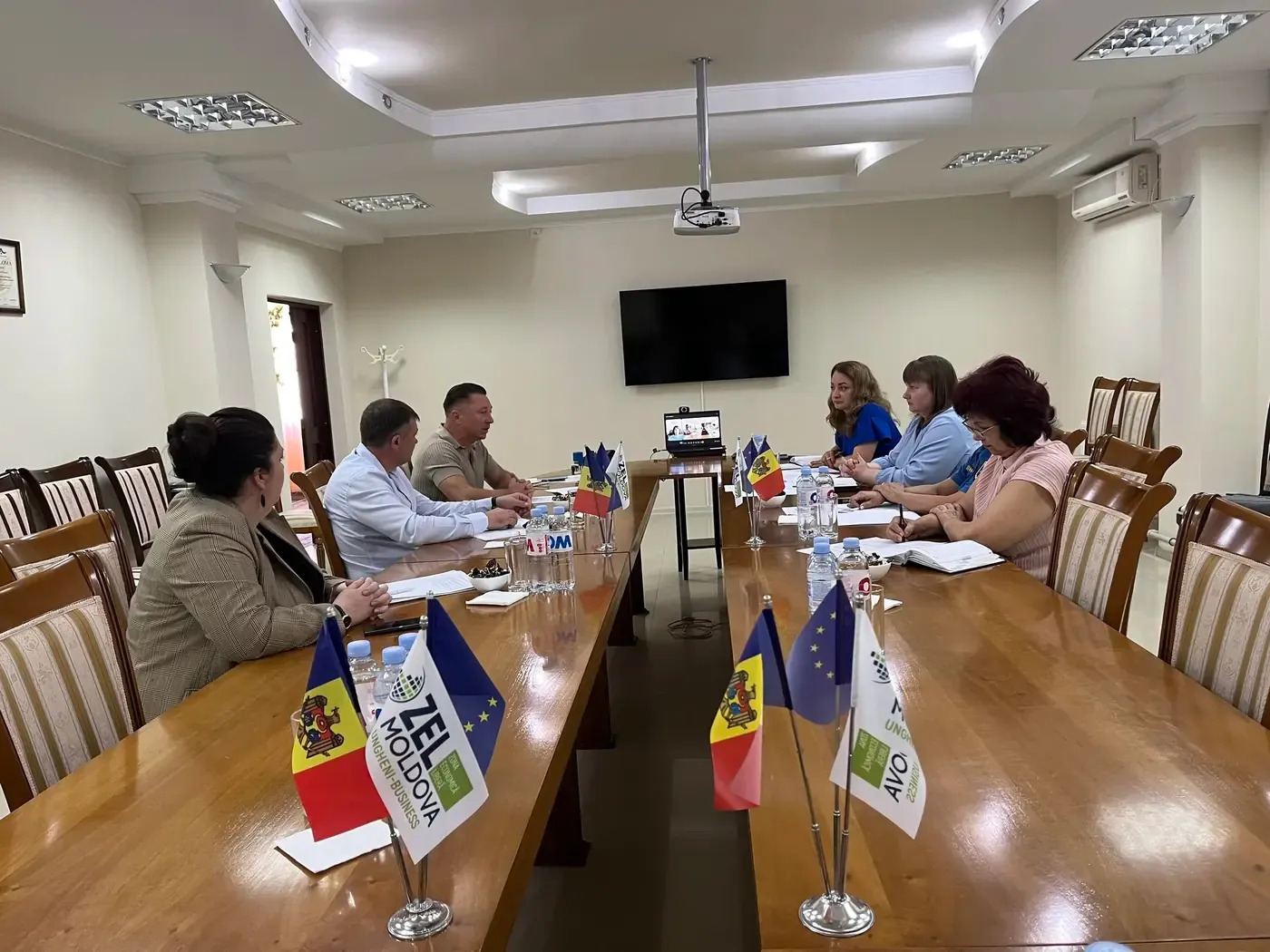 Sala de ședințe a Zonei Economice Libere Ungheni-Business, cu fanioane ZEL Moldova Ungheni-Business, ale Republicii Moldova și ale Uniunii Europene pe masă; participanții stau față în față, iar un laptop cu o videoconferință este așezat la capătul mesei