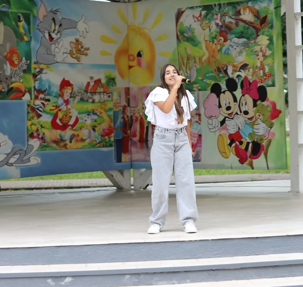 O tânără interpretă cântă la microfon pe treptele scenei din parc, în fața unei mari picturi murale cu personaje de desene animate.