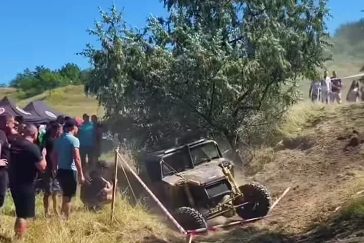 Spectatori pe marginea traseului de trial, urmărind un vehicul modificat care negociază un obstacol de teren, cu cort de echipă și dealuri verzi în fundal