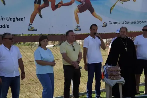 La deschiderea turneului, în fața bannerului competiției, un preot ortodox stă lângă un colac și un portret așezate pe o măsuță, alături de organizatori; unul dintre ei vorbește la microfon.