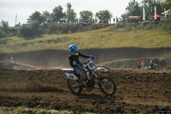 Pilot junior cu numărul 95 și cască albastră pe o motocicletă albă, pe traseul de pământ, cu mașini și spectatori în fundal.
