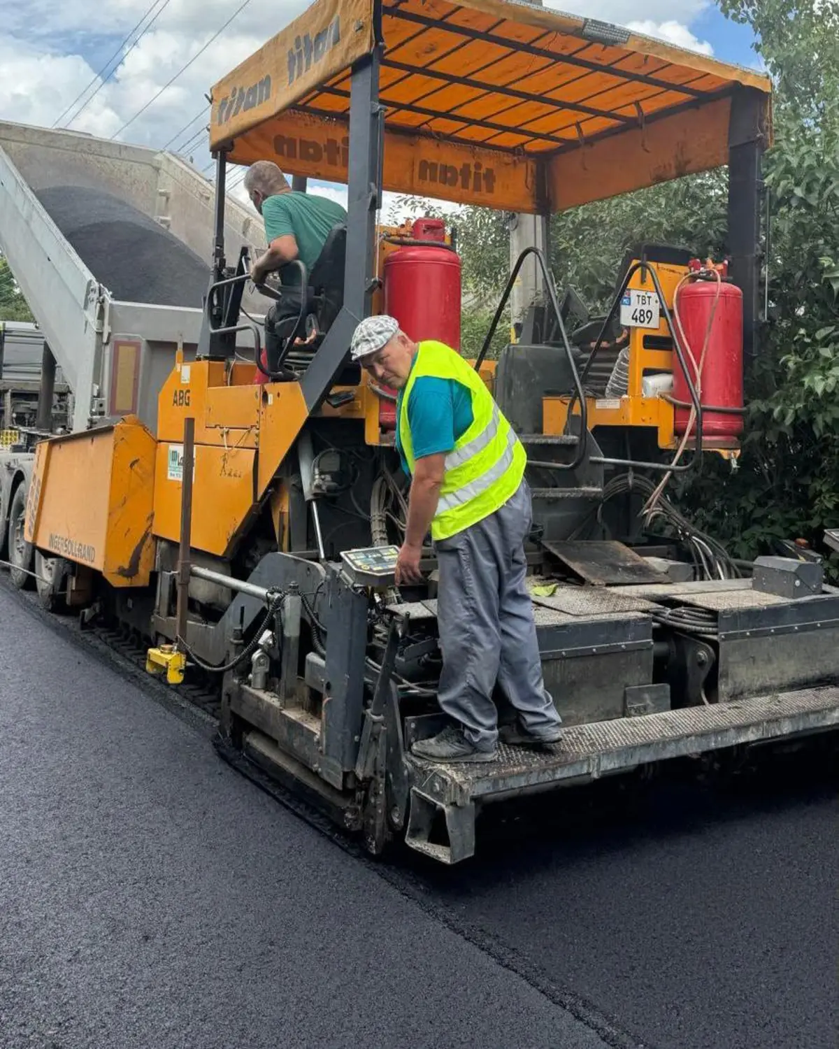 Un muncitor în vestă reflectorizantă operează comenzile finisorului de asfalt, alături de un coleg care alimentează utilajul.