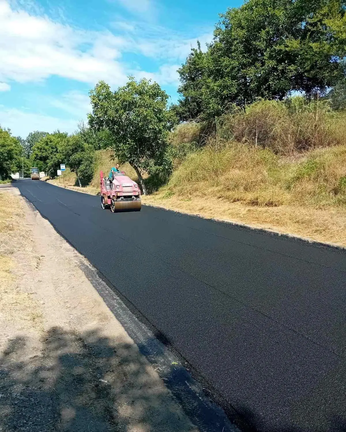 Un rulou compactor mic netezește asfaltul proaspăt pe o stradă mărginită de un indicator de stație de autobuz.