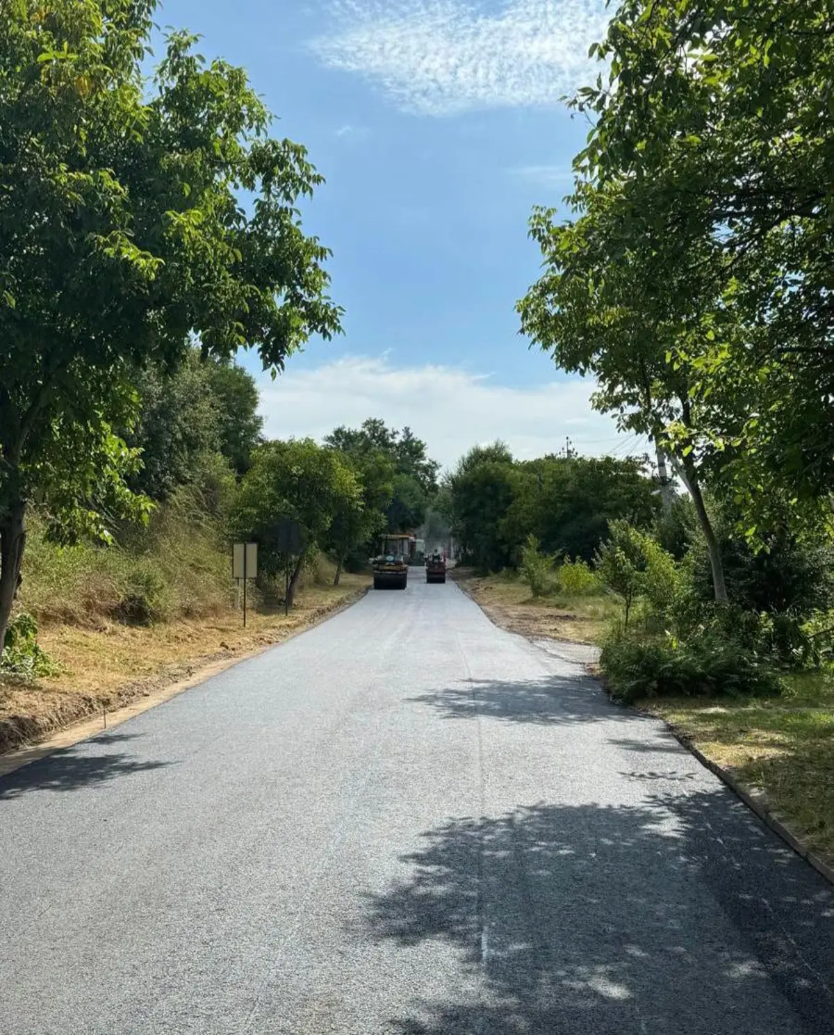 Strada proaspăt asfaltată se întinde în perspectivă printre copaci, cu utilajele de compactare la capăt.